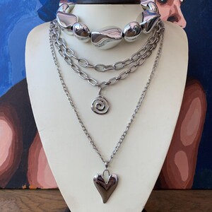 May include: A multi-strand silver-toned necklace set displayed on a white bust. The set includes a chunky choker, a chain with a spiral pendant, and a chain with a heart pendant. The necklaces have a polished, reflective finish, and are suitable for various occasions.