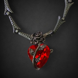 May include: A silver necklace with a heart-shaped pendant featuring a red crystal and a black rose.
