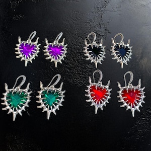 May include: Six pairs of heart-shaped earrings in various colors: purple, black, green, and red. Each earring features a silver-colored, spiky frame and a matching colored heart-shaped gem. The earrings have a hook closure.
