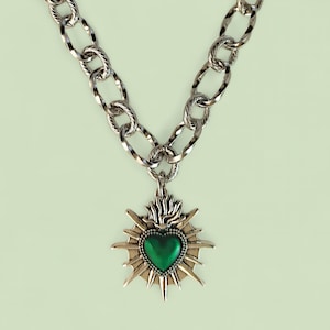 May include: Silver-toned necklace with a large, textured chain. The pendant features a green heart at the center, surrounded by a sunburst design. The necklace is a statement piece, suitable for adding a bold touch to any outfit.