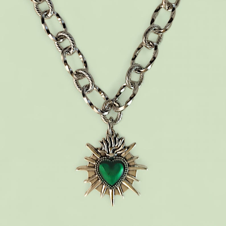May include: Silver-toned necklace with a large, textured chain. The pendant features a green heart at the center, surrounded by a sunburst design. The necklace is a statement piece, suitable for adding a bold touch to any outfit.