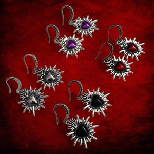 May include: Several silver earrings with heart-shaped centres in purple, red, black, and silver. Each earring has a spiky, sunburst design around the heart, and a curved hook. The earrings are displayed against a red background.