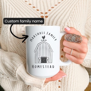 May include: White ceramic mug with a black and white barn illustration and the words "MARSHALL FAMILY HOMESTEAD". The mug also has the text "Custom family name". The mug is held by a person wearing a cream-colored sweater.