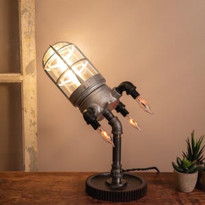 May include: Industrial-style desk lamp with a cage-like shade and exposed bulbs. The lamp features a gear-shaped base and pipe-like arms holding additional bulbs. Two small potted plants sit nearby on a wooden surface.