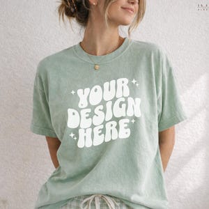 May include: A sage green t-shirt with the text "YOUR DESIGN HERE" in white, with star accents. The shirt is a relaxed fit and appears to be made of soft material. The model is wearing a gold necklace and matching plaid pants.