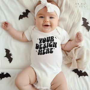 May include: White baby onesie with black text that says "YOUR DESIGN HERE" in a retro font. The onesie is on a white blanket with black bats.