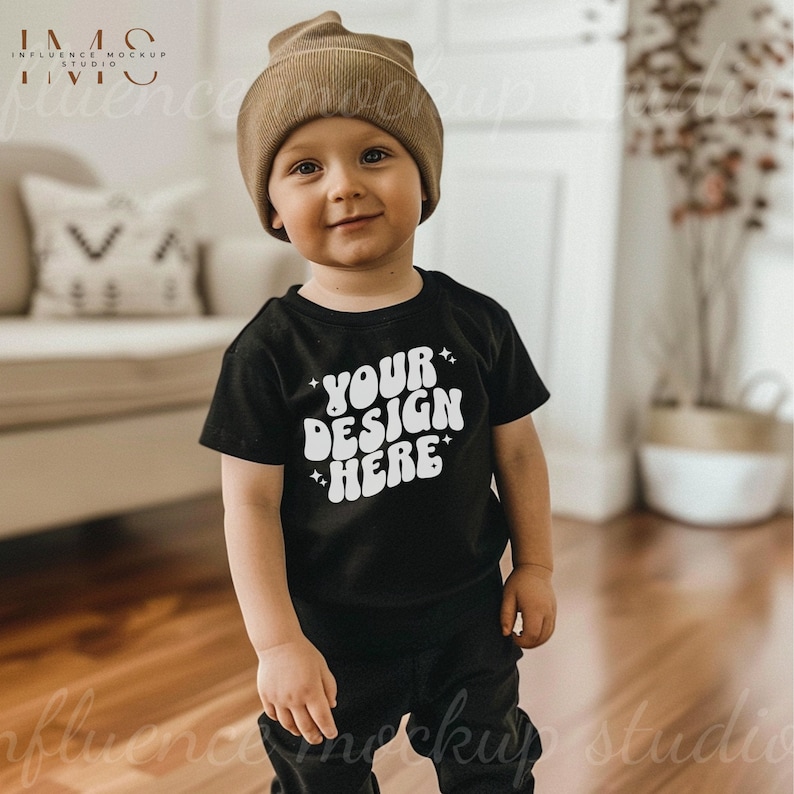 May include: A young child wearing a black t-shirt with the text "YOUR DESIGN HERE" in white. The child is also wearing a brown beanie and black pants.