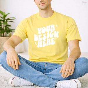 Könnte beinhalten: Ein gelbes T-Shirt mit dem weißen Text "YOUR DESIGN HERE", kombiniert mit einer blauen Jeans und weißen Turnschuhen. Die Person sitzt im Schneidersitz auf einem weißen Teppich. Eine grüne Pflanze ist im Hintergrund zu sehen.