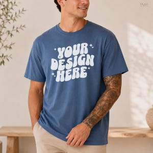 Può includere: Una maglietta blu con la scritta "YOUR DESIGN HERE" in bianco, con lettere stilizzate. La maglietta è a maniche corte e ha uno scollo tondo. Il modello indossa pantaloni color cachi.