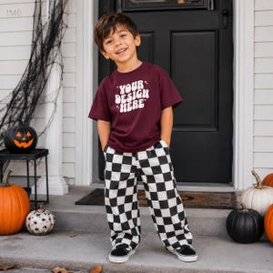 Può includere: Una t-shirt a maniche corte color bordeaux con la scritta bianca "YOUR DESIGN HERE" in un carattere ondulato, abbinata a pantaloni a scacchi bianchi e neri e scarpe da ginnastica nere. La maglietta presenta piccole stelle attorno al testo.