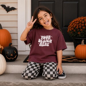 Può includere: Una t-shirt a maniche corte color bordeaux con la scritta bianca "YOUR DESIGN HERE" in un carattere ondulato di ispirazione retrò, decorata con piccole icone a forma di stella. È abbinata a pantaloni a scacchi bianchi e neri.
