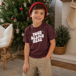 Può includere: Una t-shirt a maniche corte color marrone scuro con la scritta bianca "YOUR DESIGN HERE" in un carattere retrò ondulato con piccoli accenti a forma di stella. È abbinata a pantaloni color marrone chiaro e a un berretto rosso tinta unita.