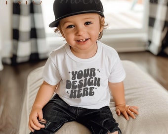 May include: A young child wearing a white t-shirt with the text "YOUR DESIGN HERE" in a retro font. The child is also wearing black jeans and a black baseball cap.