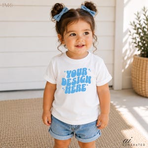 May include: A white short-sleeved t-shirt featuring the text "YOUR DESIGN HERE" in a light blue, wavy, retro-style font with small star accents. The shirt is paired with light blue denim shorts and two light blue hair bows in the hair.