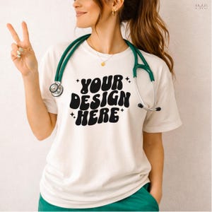 May include: Cream-colored t-shirt with the text "YOUR DESIGN HERE" in a retro font. A green stethoscope is around the neck. The person is wearing green scrubs and making a peace sign with their fingers.