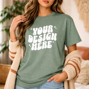 May include: Sage green t-shirt with the text "YOUR DESIGN HERE" in white, retro-style lettering. The shirt is short-sleeved and appears to be made of a soft material. A beige cardigan with brown stripes is draped over the shoulders.