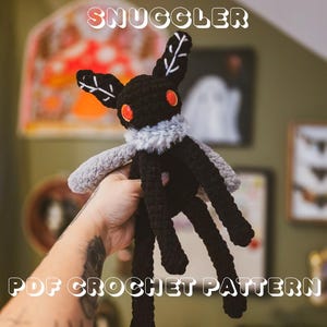 Può includere: Un peluche di Mothman all'uncinetto nero e grigio con occhi arancioni e un soffice colletto grigio. Il peluche ha gambe e ali lunghe e pendenti. Il testo "FLOPPY MOTHMAN SNUGGLER PDF CROCHET PATTERN" è visualizzato in lettere bianche.