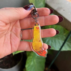 May include: A yellow chick keychain with black eyes and a red beak. The chick is made of clear plastic with a printed image inside. The keychain is attached to a silver metal ring.