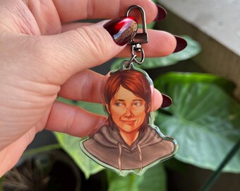 May include: A clear acrylic keychain featuring a cartoon illustration of a young woman with brown hair and brown eyes. She is wearing a gray hoodie and has a slight smile on her face.