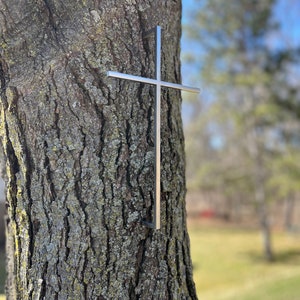 Peut inclure: Une croix en métal argenté est fixée au tronc d'un arbre. La croix est composée de deux morceaux de métal qui se croisent à angle droit. Le tronc de l'arbre est brun et a une texture rugueuse.