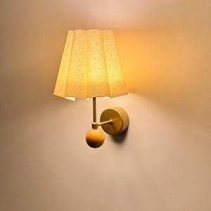May include: A wall-mounted lamp with a pleated, beige lampshade. The lamp has a gold-colored arm and a wooden sphere detail. The lamp is mounted on a beige wall, and the light from the lamp casts a warm glow.