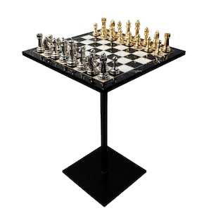 May include: A chess set with gold and silver pieces arranged on a black and white checkered board. The board is set on a square, marble-like surface, supported by a black metal stand. The stand has a square base.