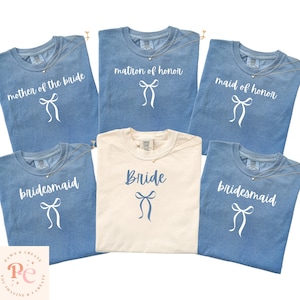 Könnte beinhalten: Eine Auswahl an blauen und cremefarbenen T-Shirts mit Text und Schleifengrafiken. Die Shirts tragen die Aufschrift "Mother of the Bride", "Matron of Honor", "Maid of Honor", "Bridesmaid" und "Bride". Jedes Shirt hat eine zarte Halskette.