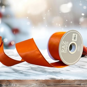 May include: A spool of orange satin ribbon with a flowing length. The ribbon is labeled "RAZ Imports" on the spool. The background is a blurred winter scene with bokeh lights and red ornaments.