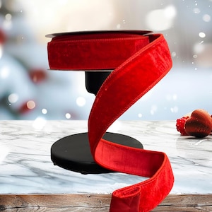 May include: A spool of vibrant red velvet ribbon, displayed on a black stand. The ribbon is unrolled, showcasing its soft texture. Decorative hearts are in the background. Perfect for crafting and holiday decor.