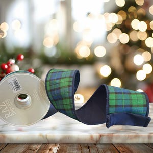 May include: A roll of wired ribbon featuring a green and navy blue plaid pattern with solid navy blue edges. The spool label displays the text RAZ Imports, 2.5 IN X 10 YDS, and 100% POLYESTER. The ribbon is unrolled to show its texture and pattern.