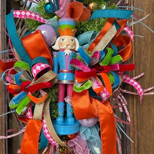 May include: A colorful Christmas wreath featuring a blue, pink, and orange nutcracker soldier. The wreath is adorned with vibrant ribbons, ornaments, and faux greenery. The design incorporates shades of teal, gold, and pink, creating a festive holiday decoration.