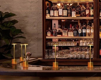May include: Four gold table lamps with a round, flat top and a cylindrical base. The lamps are on a dark brown counter top in front of a wooden bar with shelves stocked with bottles of liquor.