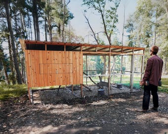 May include: A wooden chicken coop with a wire mesh enclosure. The coop has a nesting box and a covered area. A person is standing near the coop, looking at it.