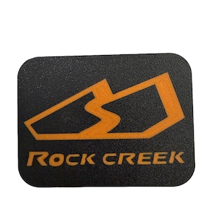 May include: A black rectangular sticker with rounded corners. The sticker features an orange graphic design and the words "ROCK CREEK" in orange block letters. The graphic appears to be a stylized mountain range.