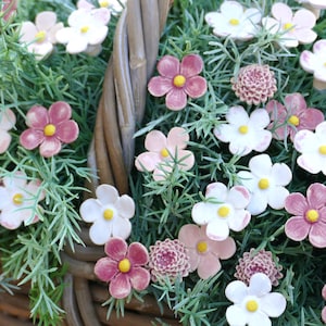 Könnte beinhalten: Ein Weidenkorb, der mit Keramikblumen in Rosa, Weiß und Lila überquillt. Die Blumen haben gelbe Zentren und sind mit grünem Laub durchsetzt. Der Korb ist aus braunem Korbgeflecht.