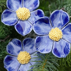Könnte beinhalten: Drei blaue Keramikblumen mit gelben Zentren. Die Blumen sind auf einem grünen Kiefernzweig angeordnet.