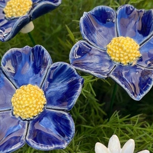 Könnte beinhalten: Handgefertigte Keramikblumen mit leuchtend blauen Blütenblättern und gelben Zentren. Die Blumen sind zwischen grünem Laub angeordnet und schaffen eine natürliche und dekorative Darstellung. Diese einzigartigen floralen Akzente verleihen jedem Raum eine künstlerische Note.