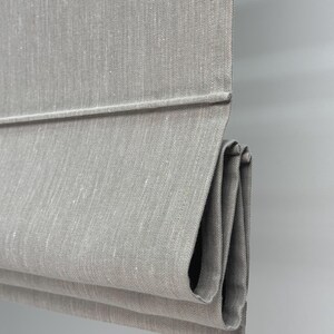 May include: Close-up of a gray Roman shade made of textured fabric. The shade is partially folded, revealing the layered construction and the fabric's subtle weave. The neutral color and simple design suggest a minimalist aesthetic.