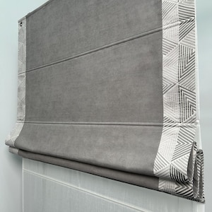 Peut inclure: Store romain gris avec une bordure à motif géométrique blanc. Le store est partiellement relevé, révélant un store en voile blanc en dessous.