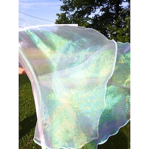 May include: A sheer, iridescent fabric in shades of white, green, and blue is held up against a backdrop of green grass and a blue sky. The fabric has a wavy, scalloped edge, and the light catches the shimmering surface.