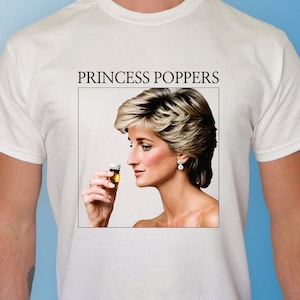 May include: White t-shirt with a graphic of Princess Diana holding a small glass of liquid. The text "Princess Poppers" is printed above the image.