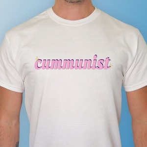 May include: A white t-shirt with the word "communist" written in pink letters with a yellow star on either side of the word.
