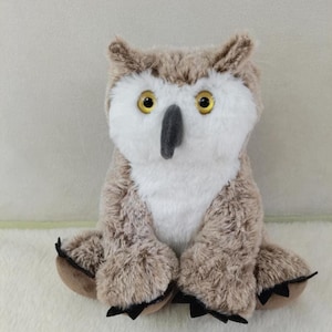 May include: A brown and white plush owl with large yellow eyes. The owl is sitting with its legs crossed and has black claws on its feet.