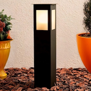 Puede incluir: Un poste de lámpara exterior de metal negro con un panel de vidrio esmerilado. El poste de la lámpara está de pie sobre un lecho de astillas de madera marrón.