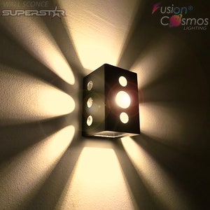 May include: A black metal wall sconce with a square shape and multiple circular cutouts. The sconce has a warm white light bulb inside, casting a warm glow.