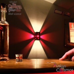 May include: A black metal wall sconce with a red light shining out from the sides. The sconce is mounted on a white wall and casts a red glow on the surrounding area. The text "Model The Cavern Bar Red Wall sconce" is visible in the top left corner of the image.