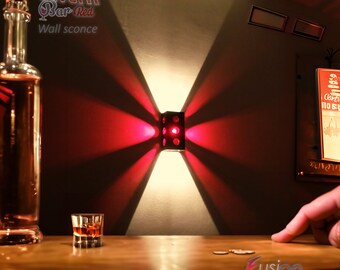 May include: A black metal wall sconce with a red light shining out from the sides. The sconce is mounted on a white wall and casts a red glow on the surrounding area. The text "Model The Cavern Bar Red Wall sconce" is visible in the top left corner of the image.