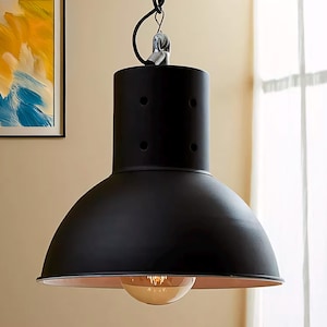 Puede incluir: Lámpara colgante negra con una pantalla redonda de metal y una bombilla transparente. La pantalla tiene una forma ligeramente abovedada y un acabado mate liso.