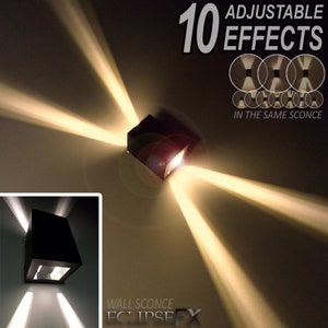Op de afbeelding: Een zwarte vierkante wandlamp met 10 instelbare lichteffecten. De lamp is gemonteerd op een witte muur en straalt lichtstralen in verschillende richtingen uit. De tekst "10 INSTELLBARE EFFECTEN" en "IN DEZELFDE WANDLAMP" is zichtbaar op de afbeelding. De tekst "WANDLAMP ECLIPSEFX" is ook zichtbaar op de afbeelding.