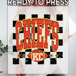 May include: Red and yellow "CHIEFS" text with a distressed look on a black and white checkered background. A red and yellow football with "KC" on it is below the text.